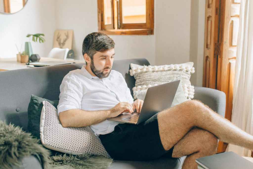 Photo of a bearded man sitting on a sofa on his laptop. He's casually reclining with the laptop balanced on his knee - showing that he's not being productive at working at home or in hybrid environments.